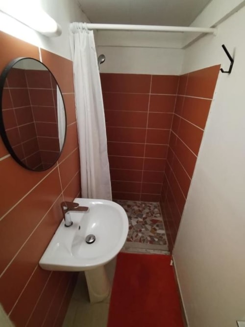 Salle de bain compacte de l'auberge de Fort-de-France avec carrelage terracotta, lavabo blanc sur piédestal, miroir rond et zone douche