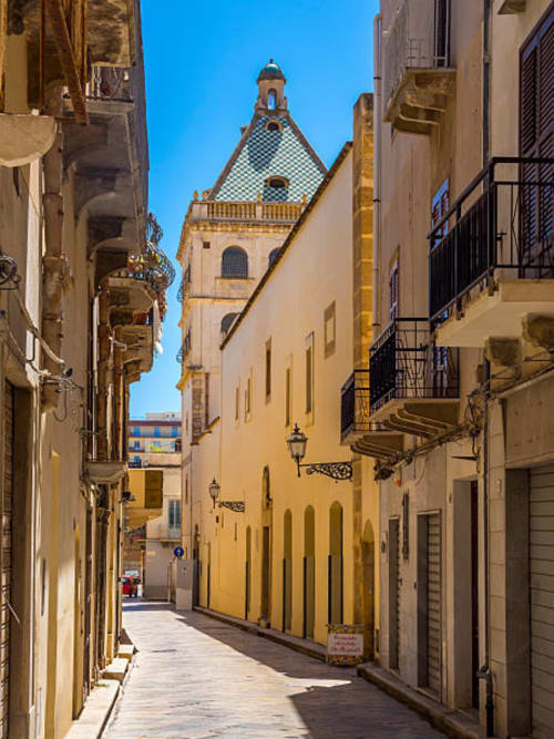Stretto vicolo in ciottolato a Marsala, Sicilia che conduce a una chiesa con cupola blu e tetto a scacchi