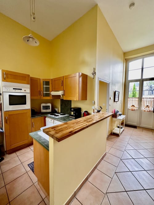 Bright yellow kitchen with wooden cabinets, island counter, and terracotta tiles in La Recreation cottage, Missillac