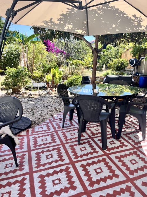 Coin repas ombragé à la Casa Monica à Cargèse avec chaises noires, tapis rouge motifs et jardin méditerranéen luxuriant