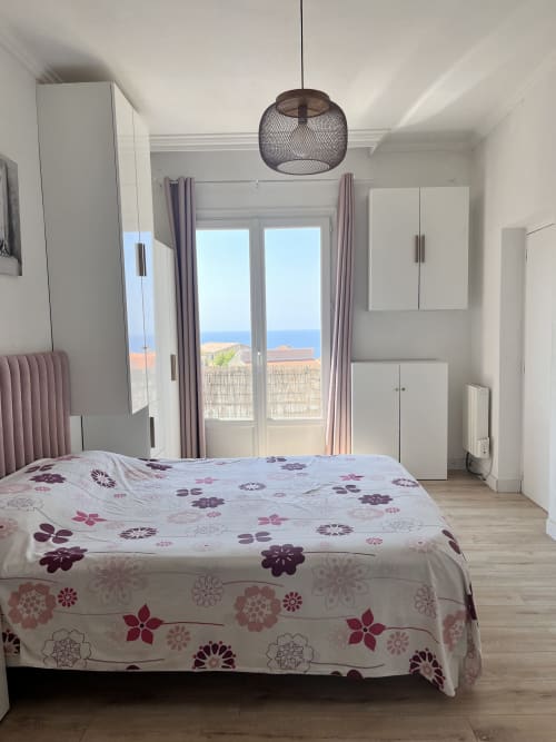Chambre lumineuse à Casa Monica à Cargèse avec vue sur la mer par les portes-fenêtres, literie fleurie, suspension moderne et blanc