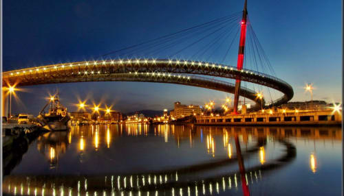Illuminated modern bridge spanning river at dusk in Pescara, Italy, with waterfront city lights reflecting in calm water