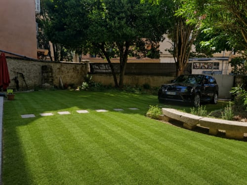 Lush green garden courtyard at Les Suites de Montigny, Aix-en-Provence, with black SUV parked, stepping stones, mature trees,