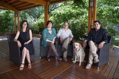 Four guests and two dogs relaxing on a wooden deck at Hotel Ristorante alle Querce in Castelbuono, Italy, surrounded by lush
