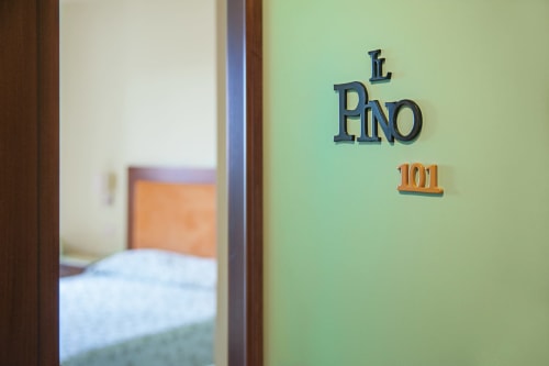 Cartello della camera Pino 101 su parete verde dell'Hotel Ristorante alle Querce, Castelbuono, con camera da letto visibile dall'ingresso