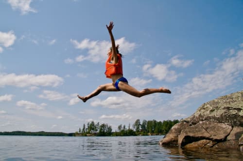 Person jumping into lake near Dolceacqua, Italy with rocky cliff and forested shoreline backdrop