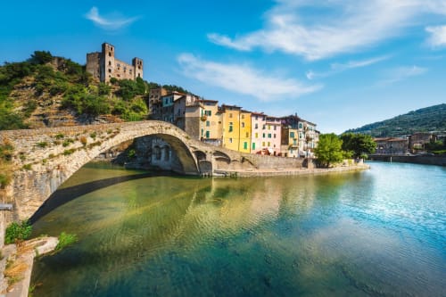 Ponte medievale in pietra sul fiume turchese a Dolceacqua, Italia, con edifici colorati e rovine del castello sulla collina