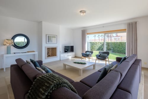 Sala de estar moderna da villa Be SilverCoast com assentos roxos, lareira branca e vista para o jardim