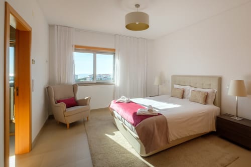 Quarto ensolarado na villa Be SilverCoast em Serra D'El Rei com cama, poltrona e vistas costeiras pelas janelas