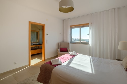 Quarto iluminado na vila Be SilverCoast em Serra D'El Rei com roupa de cama branca, porta de banheiro de madeira, poltrona e janela