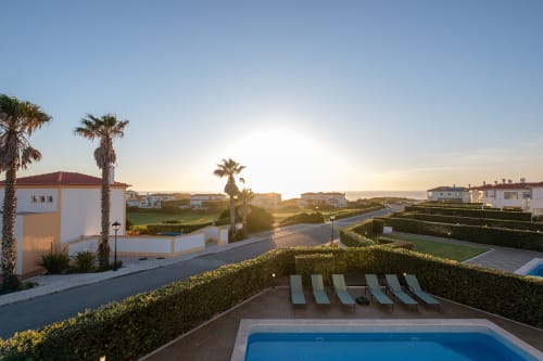 Piscina privativa e espreguiçadeiras da villa Be SilverCoast em Serra D'El Rei, Portugal ao pôr do sol com palmeiras e edifícios brancos