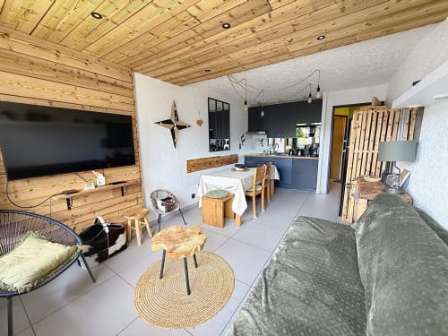 Salon d'appartement de ski moderne avec plafond en bois, TV écran plat, canapé gris, coin repas et cuisine équipée
