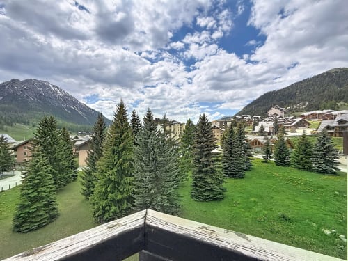 Station alpine Les Tanières de Montgenèvre avec forêts persistantes, prairie verte et pics enneigés sous ciel nuageux
