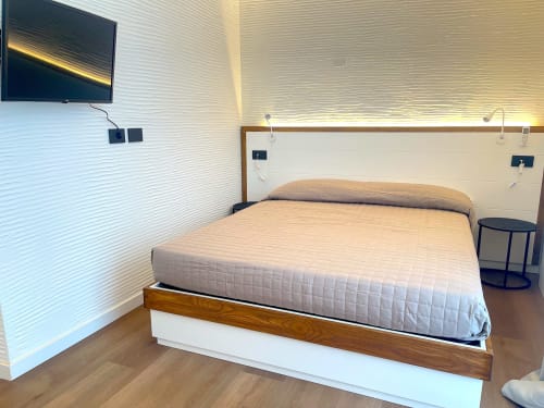 Modern hotel bedroom at Like at Home Palau with queen bed, wall-mounted TV, white textured walls, and warm lighting