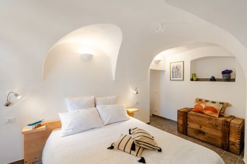 Chambre lumineuse de l'appartement Talking Stones à Dolceacqua, avec plafonds voûtés, draps blancs, banc en bois et niche de rangement.