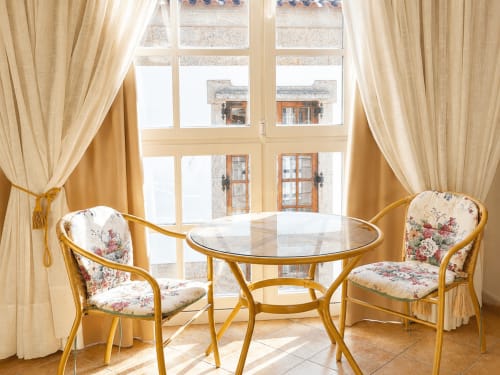 Luminosa sala de estar en Baiona con sillas doradas, mesa redonda de cristal y vistas a la ciudad a través de grandes ventanales