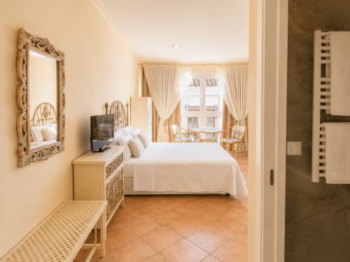 Elegante dormitorio en el apartamento Boquiera de Baiona con espejo ornamental, cama blanca, suelos de terracota y vistas a la ciudad
