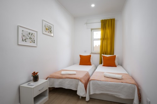Quarto duplo no hotel RB Algarve em Vilamoura com duas camas de solteiro, almofadas laranja, cortinas amarelas e comodidades modernas.