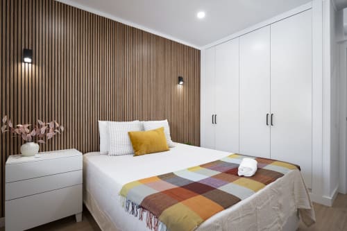 Quarto moderno da RB Algarve com parede de destaque em madeira, móveis brancos, colcha colorida e decoração minimalista