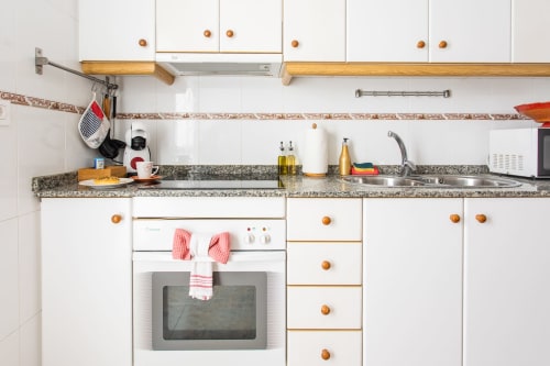 Cocina moderna con muebles blancos, tiradores de madera, encimera de granito, estufa con toalla rosa y fregadero en Haus Holiday