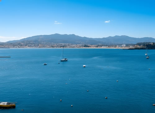 Vista de bahía turquesa en el hotel Holibai de Baiona, España con veleros y montañas