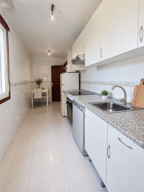 Moderna cocina de pasillo del hotel Holibai en Baiona con muebles blancos, encimera de granito y electrodomésticos de acero inoxidable