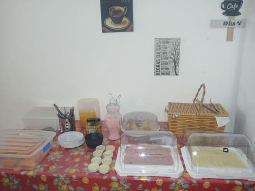 Área de refeições compartilhada do Rio Vibes Hostel com exibição de alimentos, bebidas e cesto de vime sobre toalha de mesa vermelha com estampa floral