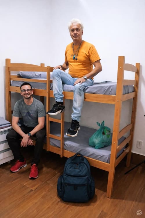 Dois hóspedes em um quarto coletivo do Rio Vibes Hostel com beliches de madeira, mochilas e piso de madeira.