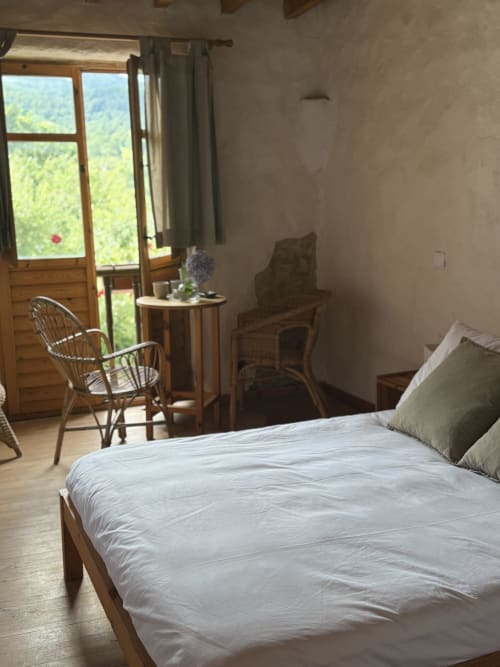 Dormitorio luminoso en Casa Aldea L'Ayalga de Infiesto con vistas a montaña, cama de madera, silla de mimbre y puertas de balcón abiertas