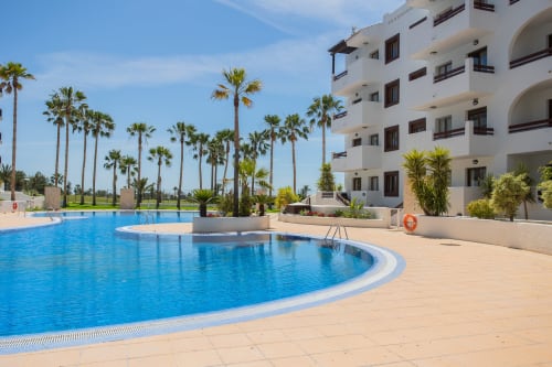 Piscina curva azul con palmeras y edificio blanco de apartamentos en Haus Holiday Homes, San Pedro del Pinatar España