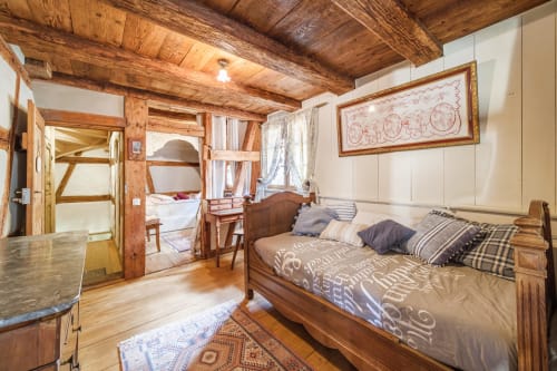 Dormitorio rústico en casita de Eguisheim con vigas de madera, cama con ropa de cama estampada, estructura de madera vista y puerta