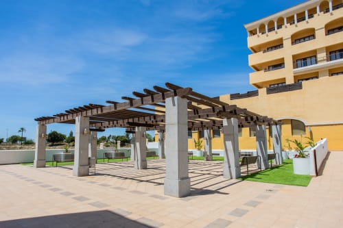 Moderna pérgola con vigas de madera en la terraza de Haus Holiday Homes, San Pedro del Pinatar, con edificio amarillo y jardín cuidado