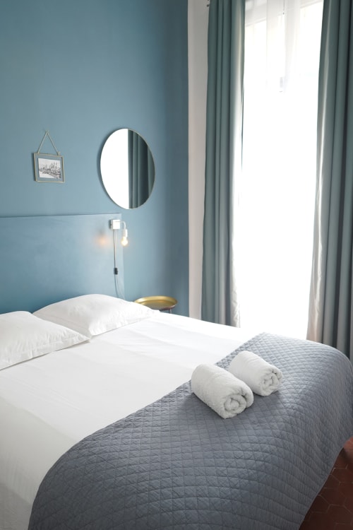 Chambre moderne à la Maison Dormoy Marseille avec murs bleus, miroir rond, oreillers blancs et serviettes roulées