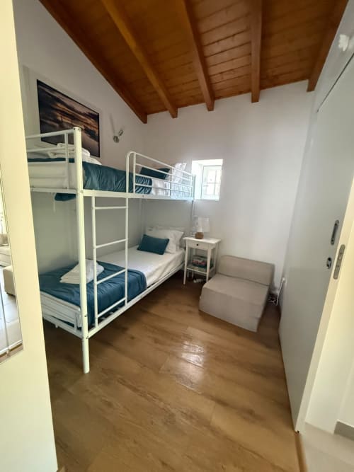 Quarto com beliches de metal branco, roupa de cama azul-piscina, teto com vigas de madeira, criado-mudo e área de estar bege