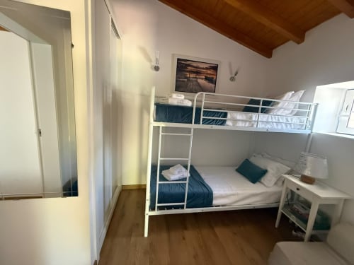 Quarto luminoso com cama beliche de metal branco, roupas de cama azul escuro, vigas de madeira, TV na parede e criado na pousada Casa Azul