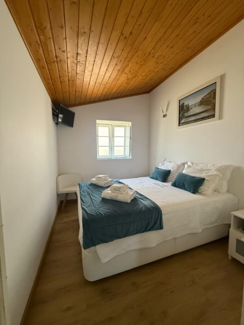 Quarto iluminado com teto de madeira, cama branca com detalhes em azul-petróleo, janela com vista para o jardim da pousada Casa Azul, Tavira