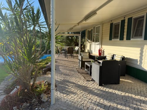 Pátio coberto com mobiliário em vime preto, plantas em vasos e vista para o bungalow Casa Verde em Monte Da Caldeirinha, Portugal