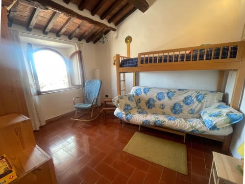 Camera rustica con travi in legno, letto a castello, divano, piastrelle in cotto e finestra ad arco 