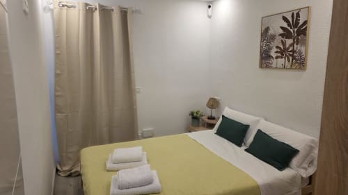 Acogedora habitación de la casita de Benicàssim con colcha amarilla, almohadas verdes, sábanas blancas y cuadros con vista al jardín