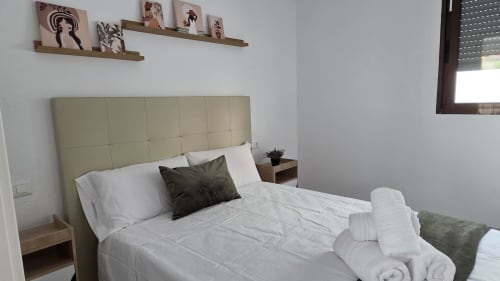 Dormitorio moderno en cabaña de Benicàssim con cabecero color crema, ropa de cama blanca, almohada oscura y mesilla de madera junto a la ventana