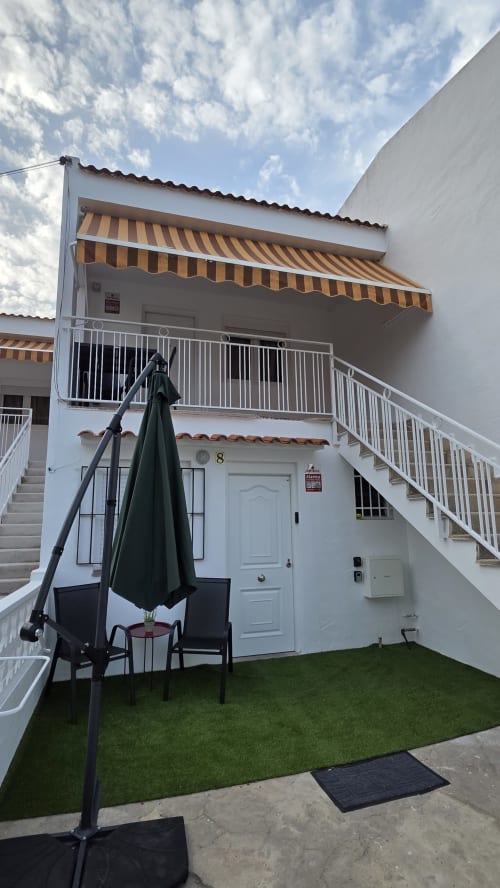 Patio ajardinado del bungalow frente al mar en Oropesa con toldo rayado, paredes blancas, sombrilla negra y césped artificial