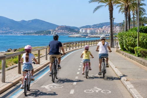 Familia disfrutando en bicicleta por el paseo costero de Baiona con palmeras y montañas al fondo