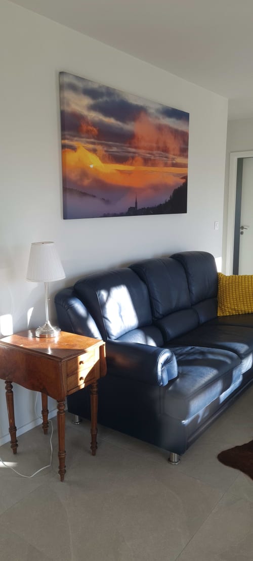 Salon moderne aux Gîtes d'Avalon avec canapé noir, table d'appoint en bois, lampe et tableau montagne coucher de soleil