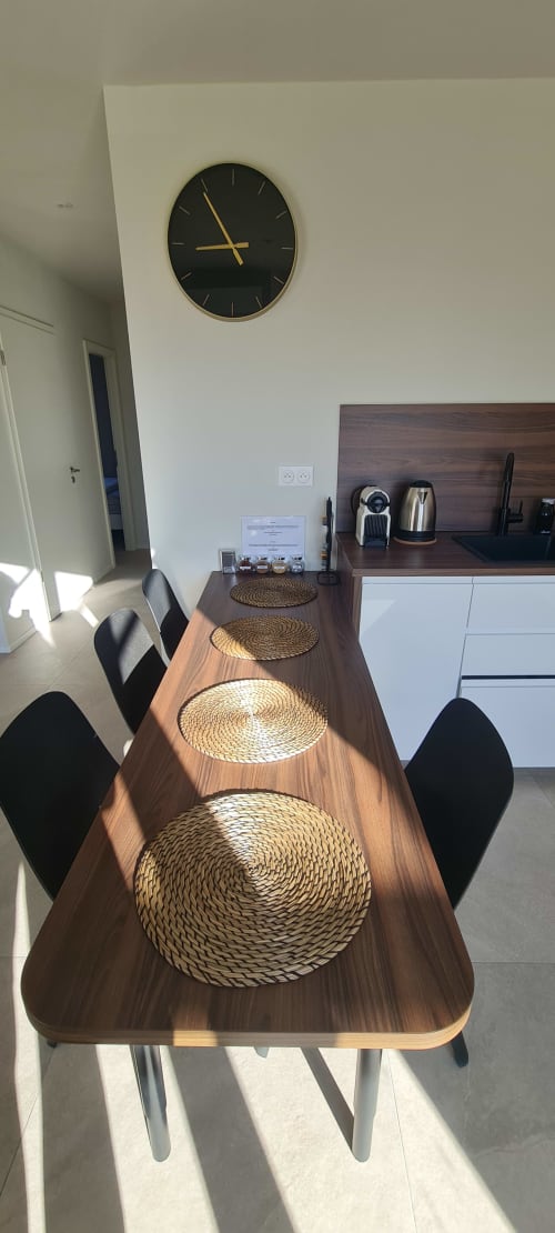 Espace salle à manger moderne à l'appartement Avalon II & III avec table en bois, sets de table tressés, chaises noires et horloge murale aux Gîtes