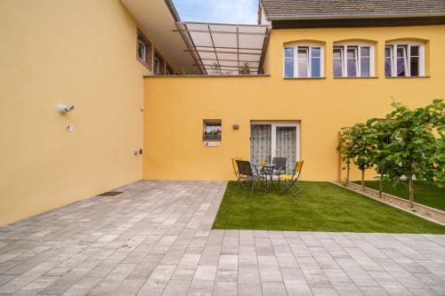 Spacious courtyard at Les Ambrades in Sigolsheim with paved terrace, lawn seating area, and yellow apartment building with