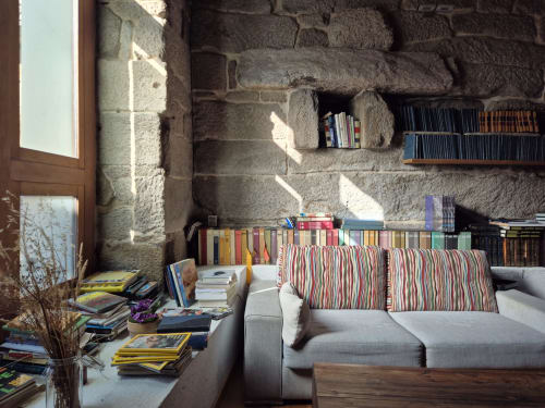 Acogedora sala de lectura con muros de piedra en Aldea Rural Pazos de Arenteiro, sofá gris, cojines a rayas y libros en estanterías
