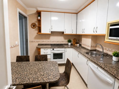Cocina moderna del hotel Holibai en Baiona con encimeras de granito, armarios blancos, mesa de comedor y electrodomésticos de acero inoxidable