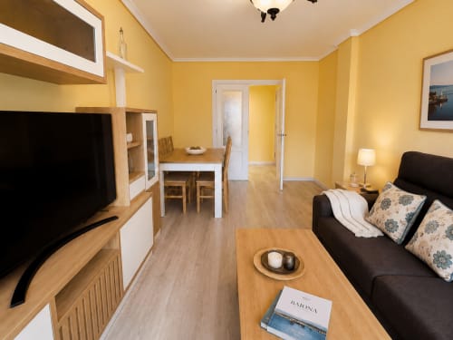 Sala de estar amarillo brillante en Holibai Baiona con sofá negro, televisor, mesa de comedor y mobiliario moderno
