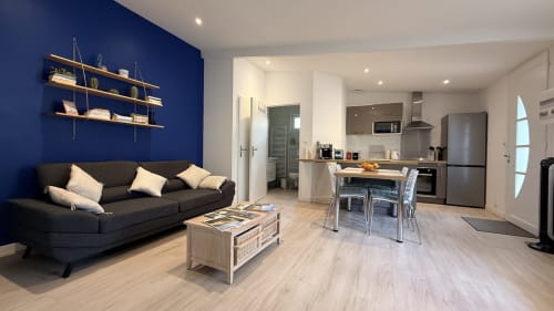 Salon de cottage moderne avec mur d'accent bleu, canapé foncé, étagères en bois, cuisine ouverte et salle à manger au Clos Bussy