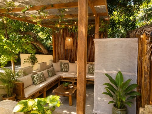 Shaded wooden pergola lounge with cream cushioned seating, patterned pillows, and lush tropical plants at Isla Mujeres